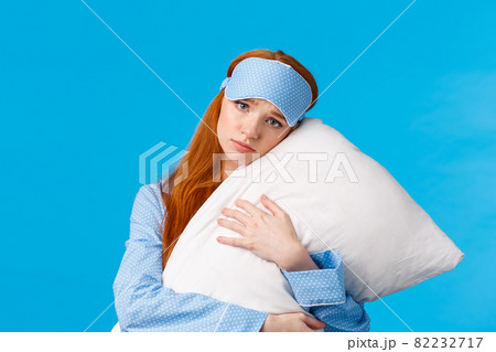 Sad redhead girl feeling depressed dont want college, lying in bed, hugging pillow, frowning and looking uneasy with concerned, nervous expression, wearing sleep mask and nightwear, blue background 82232717