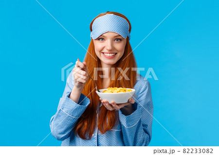 Wanna bite. Cute feminine young redhead girl in glamour nightwear, sleep mask, prepare breakfast, holding spoon and bowl, eating cereals in morning, smiling feeling energized and healthy 82233082