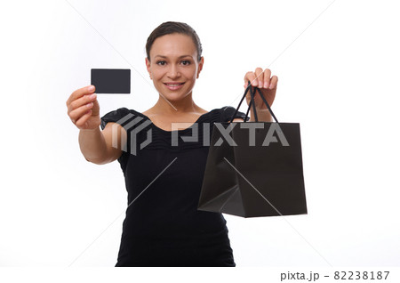 Beautiful mixed race woman looks at camera and smiles toothy smile, showing black shopping packet and plastic credit or discount card in her outstretched hands. Concept of Black Friday with copy space Beautiful mixed race woman looks at camera and smiles toothy smile, showing black shopping packet and plastic credit or discount card in her outstretched hands. Concept of Black Friday with copy space 82238187