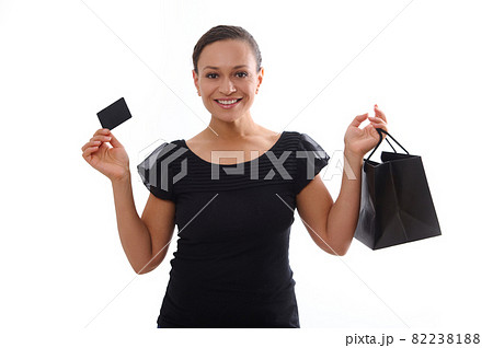 Isolated portrait on white background with copy space of attractive smiling woman dressed in black and holding a discount credit card and a shopping bag, looking at camera. Black Friday concept 82238188