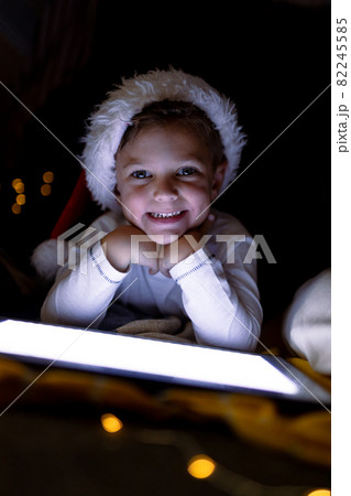Happy caucasian boy wearing santa hat, using tablet, looking at camera at christmas time 82245585