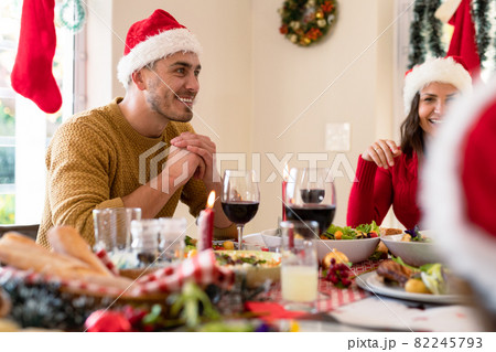 Happy caucasian couple wearing santa hats sitting at christmas table with family Happy caucasian couple wearing santa hats sitting at christmas table with family 82245793
