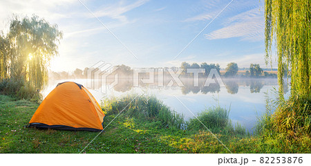 Foggy lake shore with orange tent in morning sun Foggy lake shore with orange tent in morning sun 82253876