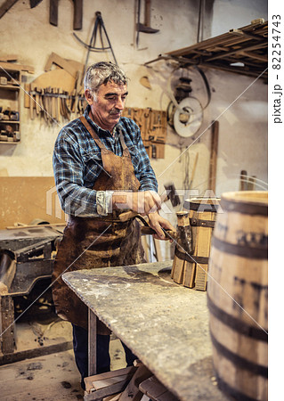 Carpenter places metal stripe on a new wooden barrel made in his cozy workshop Carpenter places metal stripe on a new wooden barrel made in his cozy workshop 82254743