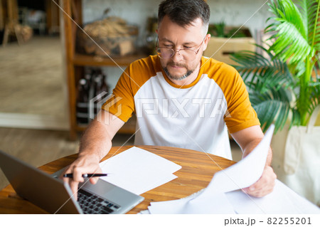 Middle-aged man works with papers at home in front of a laptop monitor. He is sitting in eyeglasses and filling online forms. Middle-aged man works with papers at home in front of a laptop monitor. He is sitting in eyeglasses and filling online forms. 82255201