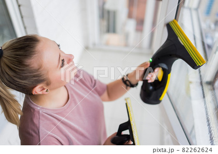 Beautiful young housewife in working clothes washing windows by professional tools at home. 82262624