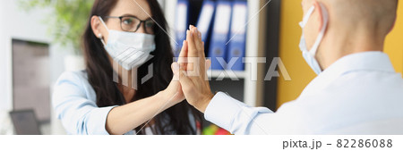 Young man and woman wearing protective medical masks giving five behind table in office Young man and woman wearing protective medical masks giving five behind table in office 82286088