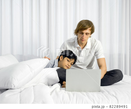 Father work at home with son. A boy napping on his father's lap while the father using laptop computer to work in the bedroom. Work life balance. 82289491