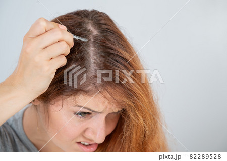 Caucasian woman finds gray hair and removes it with tweezers. Signs of aging. 82289528