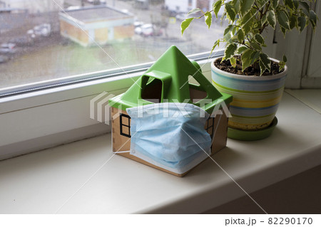 safe home concept, quarantine coronavirus. a toy house in a medical mask stands on a windowsill next to a flower in a pot safe home concept, quarantine coronavirus. a toy house in a medical mask stands on a windowsill next to a flower in a pot 82290170