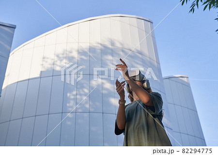 A girl is having fun in virtual reality glasses, standing against the background of a mirrored building. The concept of the future. High-quality photo 82294775