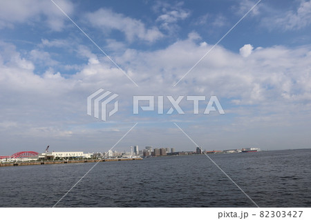 色々な色と形の雲と青空と海と赤い橋と人工島のビルの多い街と突堤とタンカーの見える風景 82303427