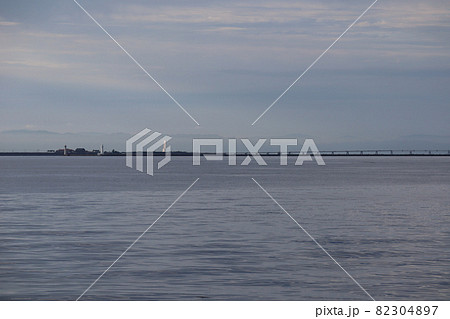 早朝の雲の多い空と海と突堤と灯台と橋と下の霞む山と白く光る高層ビルの見える風景 82304897