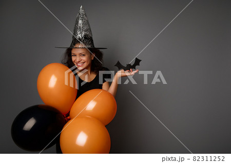 Beautiful mysterious smiling woman in wizard hat, dressed in black, holds orange black air balloons and handmade felt-cut black bat, looks at camera, poses against gray background, copy space . Beautiful mysterious smiling woman in wizard hat, dressed in black, holds orange black air balloons and handmade felt-cut black bat, looks at camera, poses against gray background, copy space . 82311252