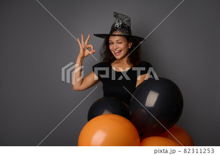 Happy joyful smiling toothy smile Hispanic woman wearing wizard hat, dressed in witch carnival costume for Halloween party, shows OK sign, poses against gray background with black balloons, copy space Happy joyful smiling toothy smile Hispanic woman wearing wizard hat, dressed in witch carnival costume for Halloween party, shows OK sign, poses against gray background with black balloons, copy space 82311253