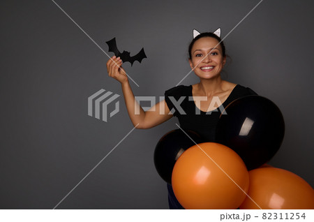Joyful Hispanic woman dressed in Halloween carnival costume looks at camera, holds handmade bat in her hand , posing with colorful black orange air balloons against gray background with copy space 82311254
