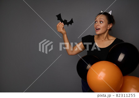 Beautiful joyful mixed race woman dressed in carnival costume looking at a felt-cut handmade bat in her hand , posing with colorful black orange air balloons against gray background with copy space Beautiful joyful mixed race woman dressed in carnival costume looking at a felt-cut handmade bat in her hand , posing with colorful black orange air balloons against gray background with copy space 82311255
