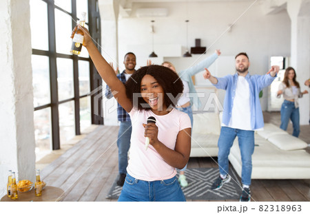 Millennial black lady with beer bottle and mic singing karaoke with diverse friends at home 82318963