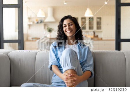 Portrait of pretty young brunette woman smiling and looking at camera, sitting on couhc at home 82319230