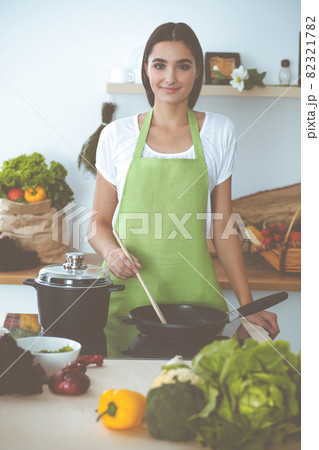 An attractive young dark-haired woman frying meal at worm pan by new keto recipe while standing and smiling in the kitchen. Cooking and householding concepts 82321782