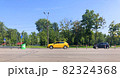 A yellow new car is driving along the road, standing out in the general traffic. A park with green trees, road signs, and a dividing strip with posts In the background. Driving school concept 82324368