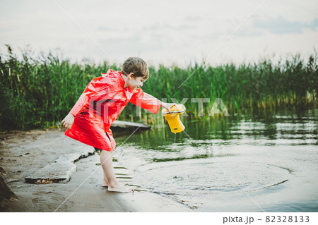 Boy in a red raincoat pours water out of yellow rubber boot into the lake. Child playing with water at pond. Kid having fun outdoors near riverside Boy in a red raincoat pours water out of yellow rubber boot into the lake. Child playing with water at pond. Kid having fun outdoors near riverside 82328133
