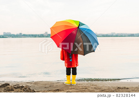 Boy in a red raincoat and yellow rubber boots holds rainbow umbrella standing at beach. Child with colourful umbrella looking at sea. School kid in a waterproof red coat holding multicolor umbrella Boy in a red raincoat and yellow rubber boots holds rainbow umbrella standing at beach. Child with colourful umbrella looking at sea. School kid in a waterproof red coat holding multicolor umbrella 82328144