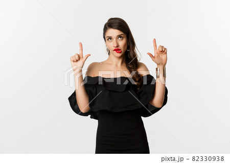 Fashion and beauty. Thoughtful attractive woman in black dress staring and pointing up, thinking with tempted look, white background 82330938