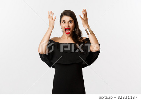 Fashion and beauty. Angry woman in black dress, shouting mad and shaking hands, grimacing outraged at camera, standing over white background Fashion and beauty. Angry woman in black dress, shouting mad and shaking hands, grimacing outraged at camera, standing over white background 82331137
