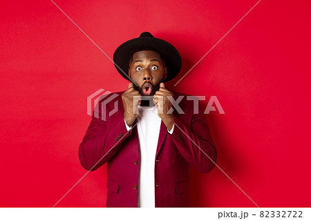 Fashion and party concept. Surprised african american male model looking shocked and amazed, saying wow and staring camera, standing against red background 82332722