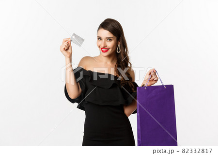 Stylish young woman in black dress going shopping, holding bag and credit card, smiling pleased, standing over white background 82332817