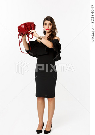 Beauty and fashion concept. Full length of sad young woman in black dress and high heels showing empty purse, sulking disappointed, standing over white background Beauty and fashion concept. Full length of sad young woman in black dress and high heels showing empty purse, sulking disappointed, standing over white background 82334047