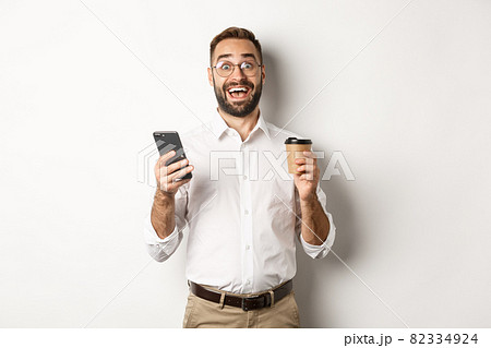 Amazed businessman drinking coffee, reacting at awesome online offer on mobile phone, standing over white background Amazed businessman drinking coffee, reacting at awesome online offer on mobile phone, standing over white background 82334924