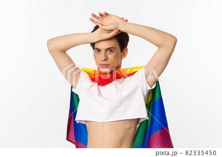 Beautiful gay man with glitter on face, wearing crop top and rainbow lgbt flag, posing against white background Beautiful gay man with glitter on face, wearing crop top and rainbow lgbt flag, posing against white background 82335450