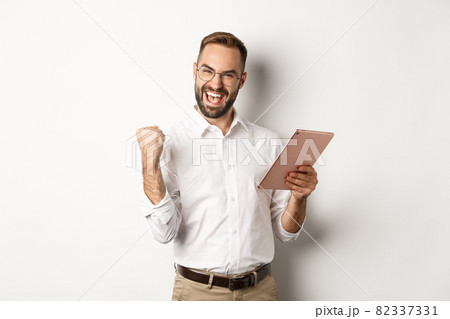 Successful businessman rejoicing on winning online, reading on digital tablet and making fist pump, triumphing, standing over white background 82337331