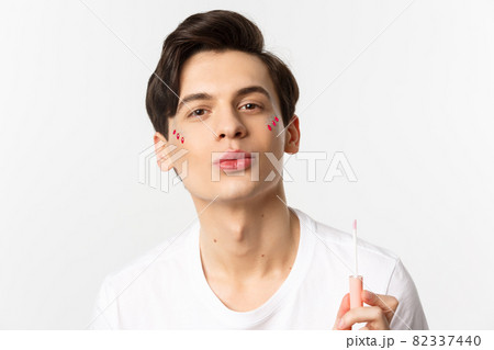 Close-up of beautiful gay man with glitter on face pucker lips after applying lip gloss, standing over white background 82337440