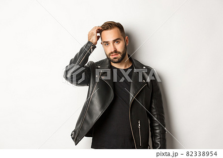Handsome macho man in black biker jacket, touching his haircut and looking cool, white background 82338954