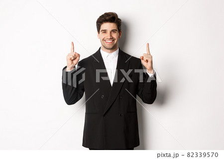Portrait of attractive caucasian man in stylish black suit, pointing fingers up and smiling, showing christmas advertisement, standing over white background Portrait of attractive caucasian man in stylish black suit, pointing fingers up and smiling, showing christmas advertisement, standing over white background 82339570