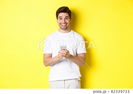 Man smiling and looking happy after reading promo offer on smartphone, standing against yellow background in white t-shirt 82339733