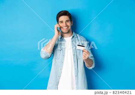Handsome man calling bank and holding credit card, having mobile conversation, standing over blue background Handsome man calling bank and holding credit card, having mobile conversation, standing over blue background 82340421