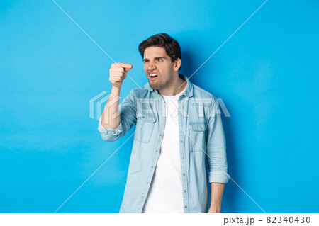 Confused guy holding something tiny in fingers and looking at it puzzled, standing over blue background 82340430
