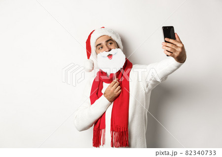 Christmas party and celebration concept. Young man taking selfie with funny white beard Santa mask and hat, posing for photo on mobile phone, studio background 82340733