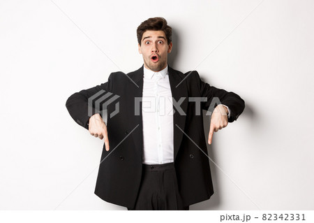 Portrait of surprised and excited man in black suit, pointing fingers down and showing christmas advertisement, standing over white background Portrait of surprised and excited man in black suit, pointing fingers down and showing christmas advertisement, standing over white background 82342331