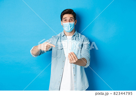 Concept of coronavirus, quarantine and social distancing. Young man pointing at medical masks, preventing measures from covid-19, standing over blue background 82344795