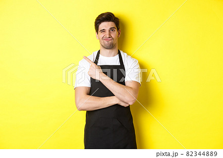 Displeased barista in black apron pointing finger at upper left corner, grimacing skeptical, dislike something, standing against yellow background 82344889