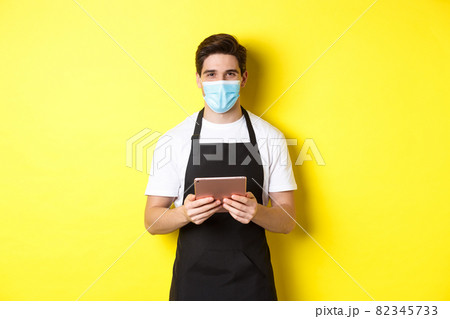 Concept of covid-19, small business and pandemic. Waiter in black apron and medical mask taking order, holding digital tablet, standing over yellow background Concept of covid-19, small business and pandemic. Waiter in black apron and medical mask taking order, holding digital tablet, standing over yellow background 82345733