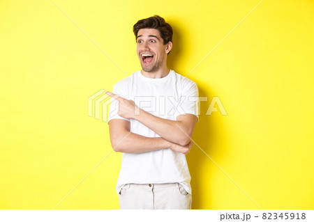 Excited smiling man pointing and looking left, checking out promo offer, standing over yellow background 82345918