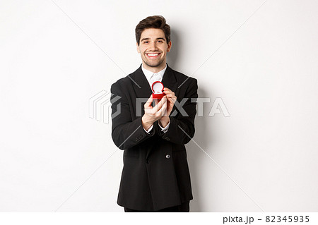 Image of handsome boyfriend in black suit making a proposal, asking to marry him and showing wedding ring, standing over white background Image of handsome boyfriend in black suit making a proposal, asking to marry him and showing wedding ring, standing over white background 82345935