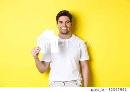 Real estate concept. Young man in white t-shirt holding paper house model and smiling, searching for apartment, standing over yellow background Real estate concept. Young man in white t-shirt holding paper house model and smiling, searching for apartment, standing over yellow background 82345941