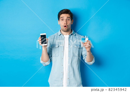 Man looking surprised, showing smartphone screen and glass of water, standing over blue background Man looking surprised, showing smartphone screen and glass of water, standing over blue background 82345942